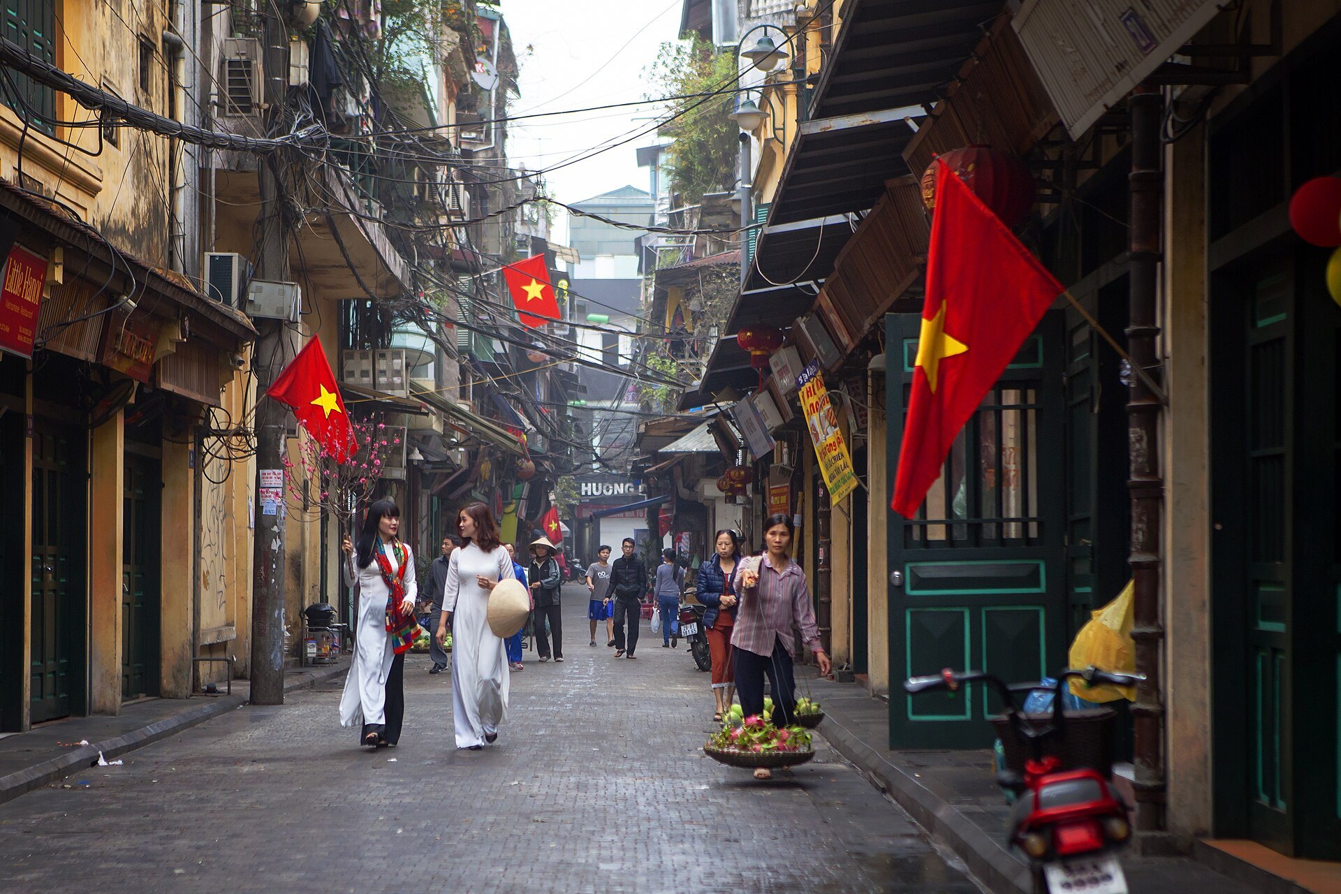Tạ Hiện street in the Old Quarter — ochre shophouses down both sides, red Vietnamese flags hanging overhead, and women in white áo dài walking between them