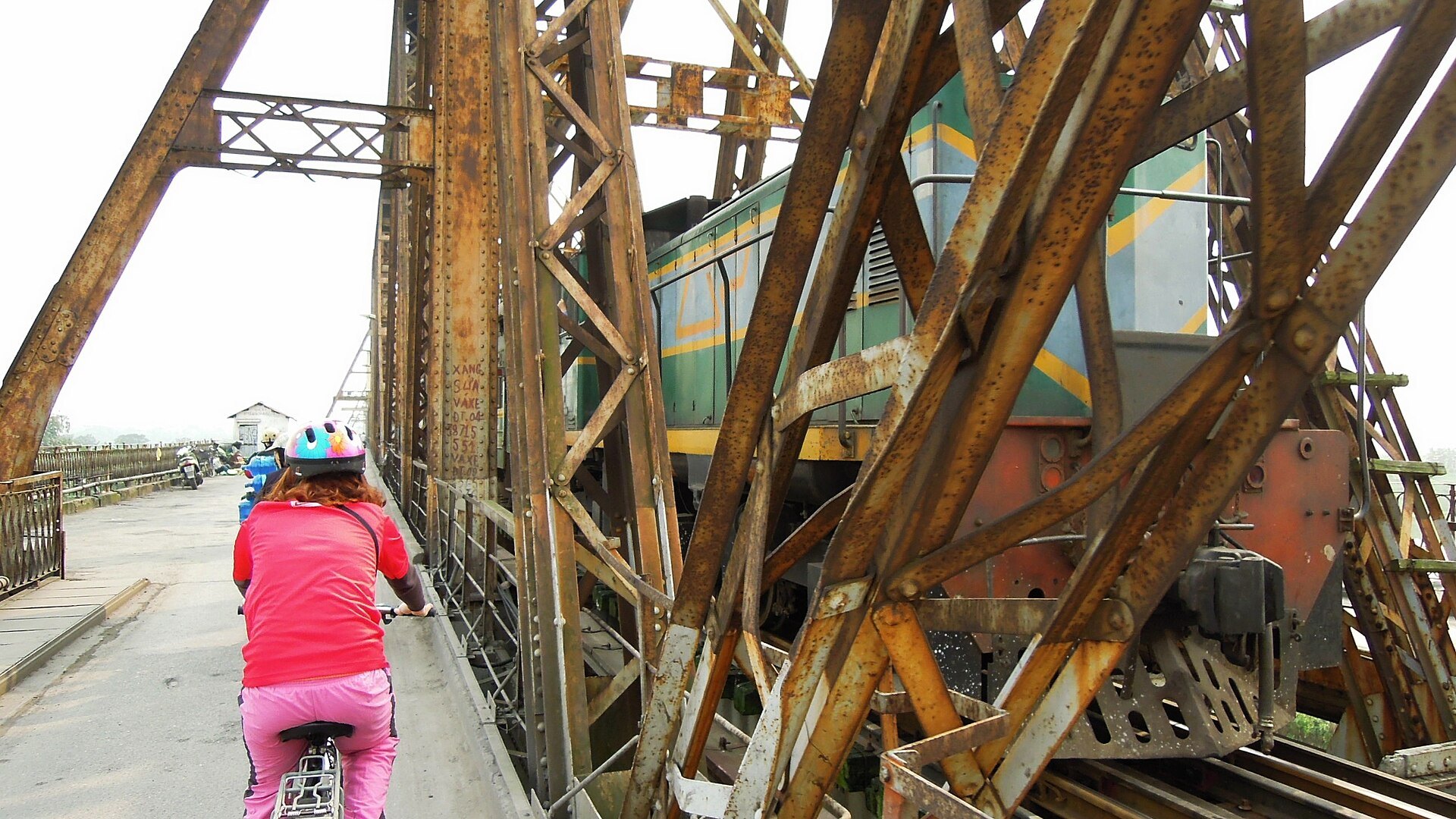 A train crossing Long Bien Bridge with a cyclist on the walkway beside it